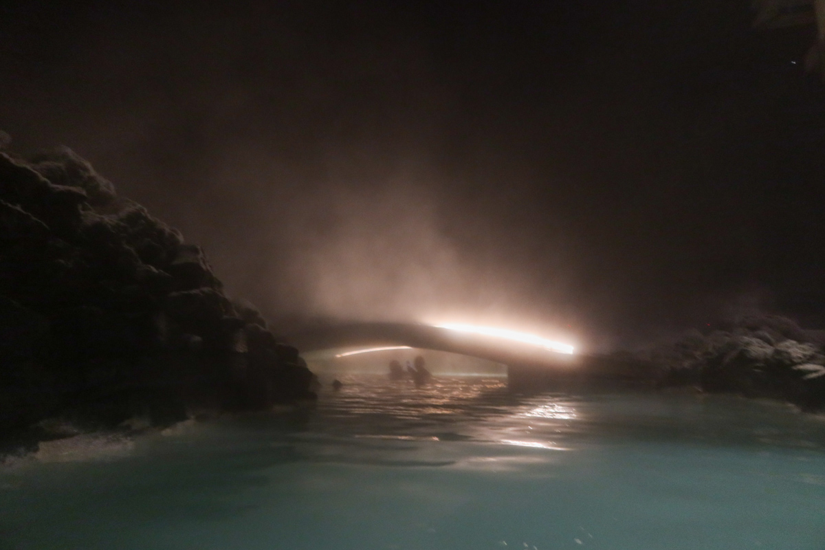 Blue water of the lagoon in the bottom of the shot, with steam rising into the dark sky at the top of the photo. 