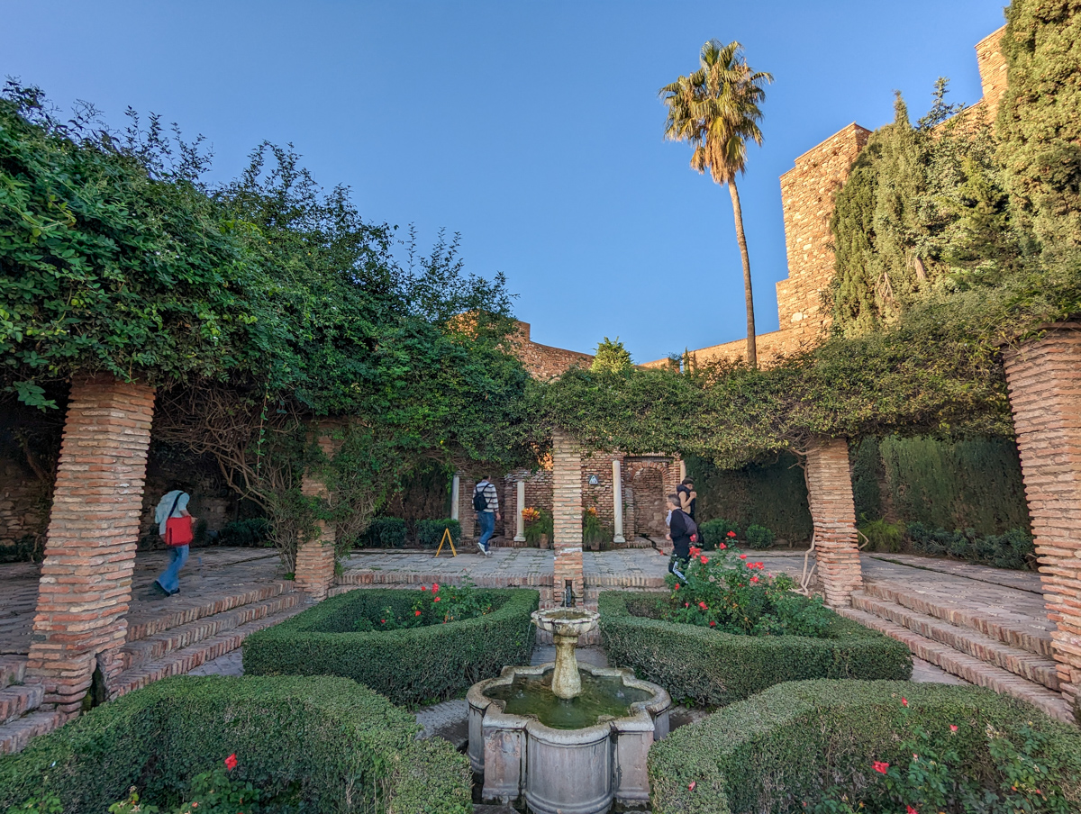 Gardens in the Alcazaba, with pillars around the side of a bush complex and palm trees in the background. 